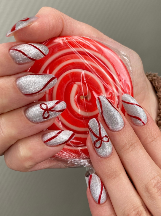 Новогодний маникюр 2026 Архангельск: Праздничный дизайн на миндалевидных ногтях с серебристым глиттерным покрытием и росписью красными ленточками/бантами, имитирующей карамель. Идеи праздничного маникюра для встречи Нового года